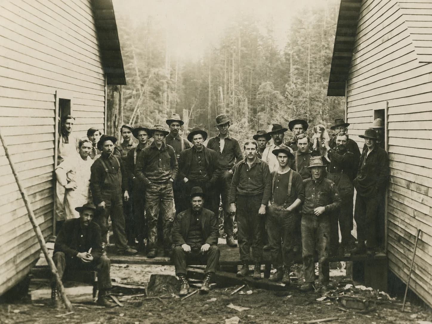 Large group of lumber camp workers gathered between two wooden bunkhouses