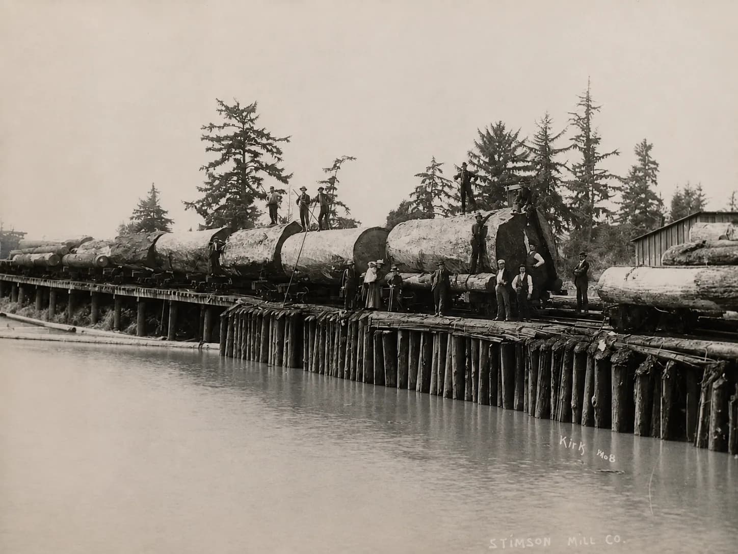 Stimson Mill Company workers standing on enormous logs beside a riverside railroad